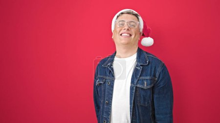 Téléchargez les photos : Jeune homme caucasien souriant confiant portant un chapeau de Noël sur fond rouge isolé - en image libre de droit