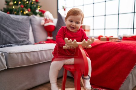 Photo for Adorable redhead toddler playing with reindeer rocking by christmas tree at home - Royalty Free Image
