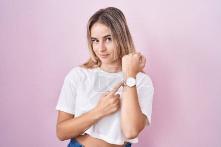Téléchargez les photos : Young blonde woman standing over pink background in hurry pointing to watch time, impatience, looking at the camera with relaxed expression - en image libre de droit