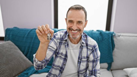 Téléchargez les photos : Homme d'âge moyen aux cheveux gris souriant, tenant la clé en toute confiance lorsqu'il est assis sur un canapé, rayonnant de positivité dans sa nouvelle maison, profitant du bonheur d'un investissement immobilier réussi. - en image libre de droit
