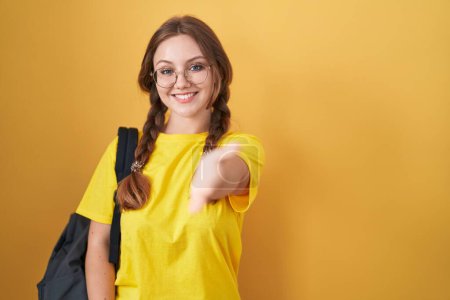 Photo for Young caucasian woman wearing student backpack over yellow background smiling friendly offering handshake as greeting and welcoming. successful business. - Royalty Free Image
