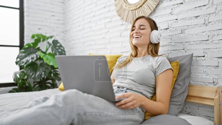 Téléchargez les photos : Jolie jeune femme blonde se détendre sur le lit dans une chambre confortable, profiter de la musique du matin à travers des écouteurs tout en explorant le monde en ligne sur ordinateur portable. - en image libre de droit