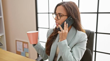 Photo for Young, beautiful, and boss! hispanic business woman engrossed in serious cellphone conversation over a relaxing cup of coffee in the office - Royalty Free Image