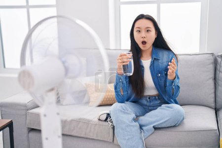 Foto de Joven mujer asiática bebiendo vaso de agua disfrutando del aire de ventilador asustado y sorprendido con la boca abierta para sorpresa, cara de incredulidad - Imagen libre de derechos