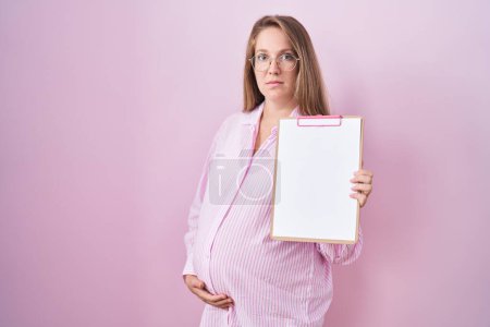Photo for Young pregnant woman holding clipboard relaxed with serious expression on face. simple and natural looking at the camera. - Royalty Free Image