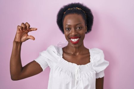 Téléchargez les photos : Femme africaine avec les cheveux bouclés debout sur fond rose geste souriant et confiant avec la main faisant signe de petite taille avec les doigts à la recherche et la caméra. concept de mesure. - en image libre de droit
