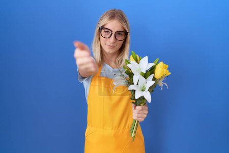 Foto de Mujer joven caucásica con delantal florista sosteniendo flores sonrientes amistosas ofreciendo apretón de manos como saludo y bienvenida. negocios exitosos. - Imagen libre de derechos