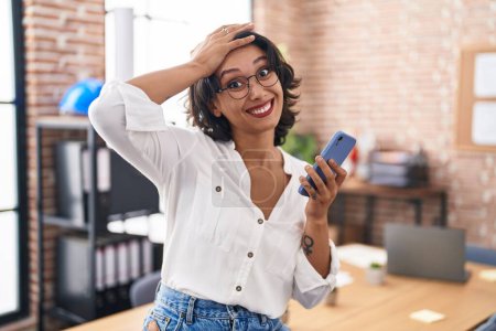 Téléchargez les photos : Jeune femme hispanique travaillant au bureau à l'aide d'un smartphone stressé et frustré avec la main sur la tête, visage surpris et en colère - en image libre de droit