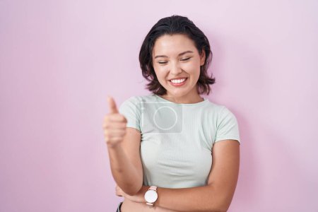 Téléchargez les photos : Jeune femme hispanique debout sur fond rose regardant fier, souriant faire les pouces vers le haut geste sur le côté - en image libre de droit