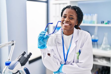 Foto de Científica afroamericana sonriendo confiada sosteniendo gafas de seguridad en laboratorio - Imagen libre de derechos