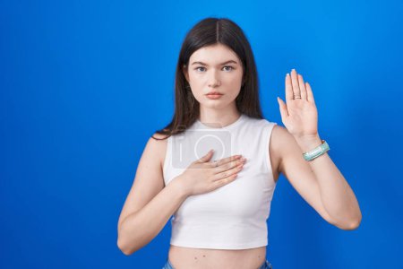 Téléchargez les photos : Jeune femme caucasienne debout sur fond bleu jurant avec la main sur la poitrine et la paume ouverte, faire un serment de fidélité promesse - en image libre de droit