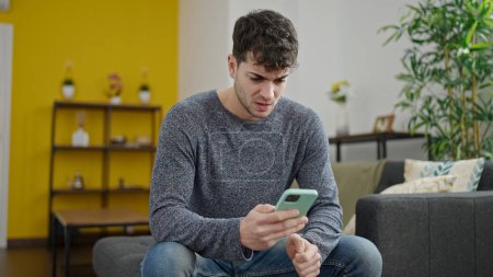 Téléchargez les photos : Jeune homme hispanique utilisant smartphone inquiet assis sur le canapé à la maison - en image libre de droit