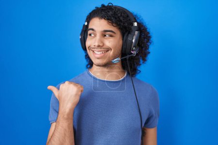 Téléchargez les photos : Homme hispanique aux cheveux bouclés écoutant de la musique à l'aide d'écouteurs souriant avec un visage heureux regardant et pointant vers le côté avec le pouce vers le haut. - en image libre de droit