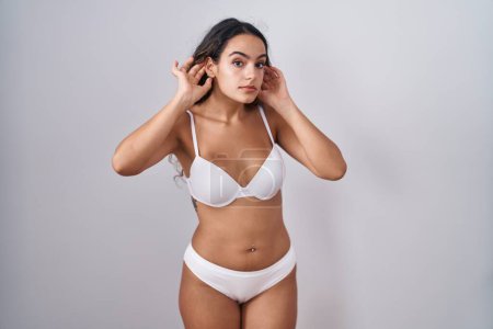 Téléchargez les photos : Jeune femme hispanique portant de la lingerie blanche essayant d'entendre les deux mains sur le geste de l'oreille, curieuse de commérages. problème auditif, sourd - en image libre de droit