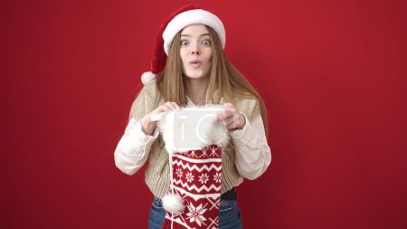 Photo for Young blonde woman wearing christmas hat looking inside of sock over isolated red background - Royalty Free Image
