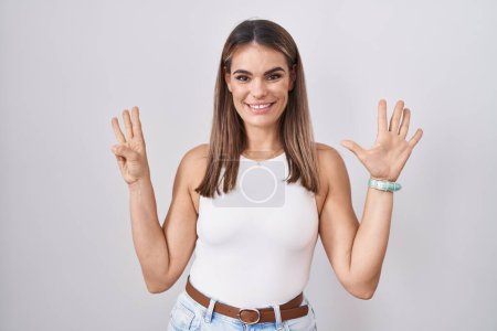 Foto de Mujer joven hispana de pie sobre fondo blanco mostrando y señalando hacia arriba con los dedos número ocho mientras sonríe confiada y feliz. - Imagen libre de derechos