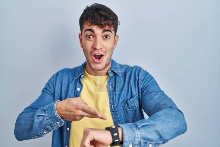 Photo for Young hispanic man standing over blue background in hurry pointing to watch time, impatience, upset and angry for deadline delay - Royalty Free Image