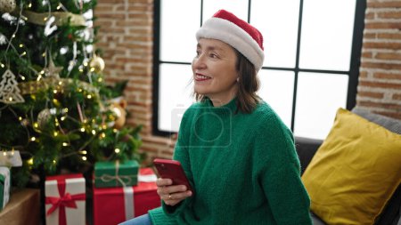 Téléchargez les photos : Femme hispanique mature aux cheveux gris utilisant un smartphone assis sur le canapé près de l'arbre de Noël à la maison - en image libre de droit