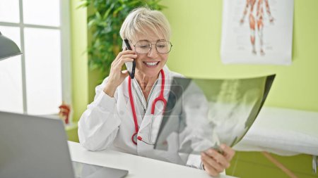 Foto de Edad media rubia mujer médico hablando en el teléfono inteligente celebración de rayos X en la clínica - Imagen libre de derechos
