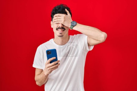 Téléchargez les photos : Homme hispanique utilisant smartphone sur fond rouge souriant et riant avec la main sur le visage couvrant les yeux pour la surprise. concept aveugle. - en image libre de droit
