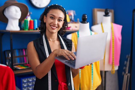 Photo for Young beautiful hispanic woman tailor smiling confident using laptop at clothing factory - Royalty Free Image