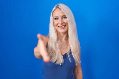 Foto de Mujer caucásica de pie sobre fondo azul sonriendo amistoso ofreciendo apretón de manos como saludo y bienvenida. negocios exitosos. - Imagen libre de derechos