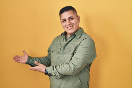 Foto de Joven hispano de pie sobre fondo amarillo invitando a entrar sonriente natural con la mano abierta - Imagen libre de derechos