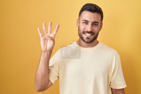 Téléchargez les photos : Bel homme hispanique debout sur fond jaune montrant et pointant vers le haut avec les doigts numéro quatre tout en souriant confiant et heureux. - en image libre de droit