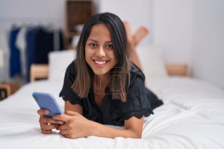 Foto de Young hispanic woman using smartphone lying on bed at bedroom - Imagen libre de derechos