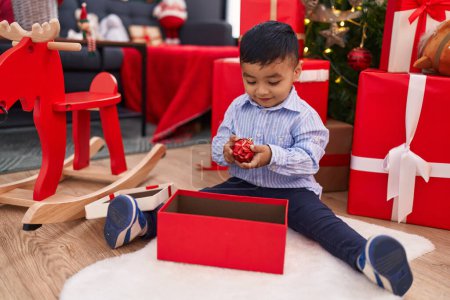 Foto de Adorable niño hispano sosteniendo la decoración de la bola de Navidad sentado en el suelo en casa - Imagen libre de derechos