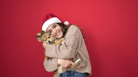 Téléchargez les photos : Jeune femme hispanique avec chien souriant confiant portant un chapeau de Noël sur fond rouge isolé - en image libre de droit