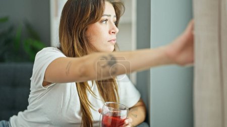 Téléchargez les photos : Jeune belle femme hispanique boire du thé regarder jeter la fenêtre avec un visage sérieux à la maison - en image libre de droit