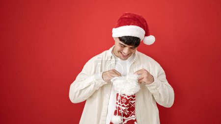 Photo for Young hispanic man wearing christmas hat looking inside of sock over isolated red background - Royalty Free Image