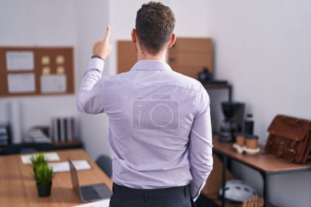 Téléchargez les photos : Jeune homme hispanique au bureau posant vers l'arrière pointant vers l'avant avec la main du doigt - en image libre de droit