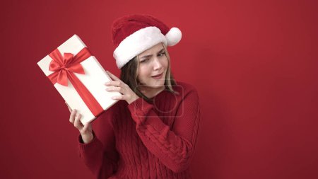 Foto de Mujer rubia joven con sombrero de Navidad escuchar sonido regalo sobre fondo rojo aislado - Imagen libre de derechos