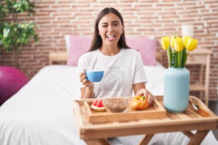 Foto de Joven mujer hispana comiendo pasteles para desayunar sentada en la cama sacando la lengua feliz con expresión divertida. - Imagen libre de derechos