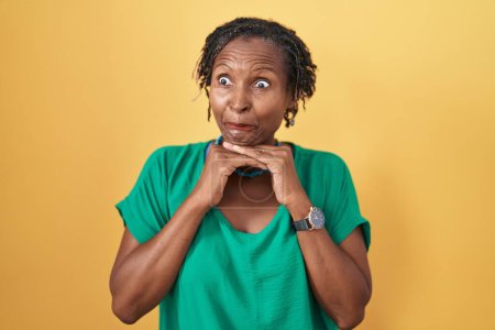 Foto de Mujer africana con rastas de pie sobre fondo amarillo riendo nerviosa y emocionada con las manos en la barbilla mirando a un lado - Imagen libre de derechos