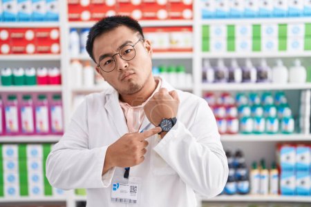 Téléchargez les photos : Jeune homme chinois travaillant à la pharmacie pharmacie pressé pointant vers regarder le temps, l'impatience, regarder la caméra avec une expression détendue - en image libre de droit