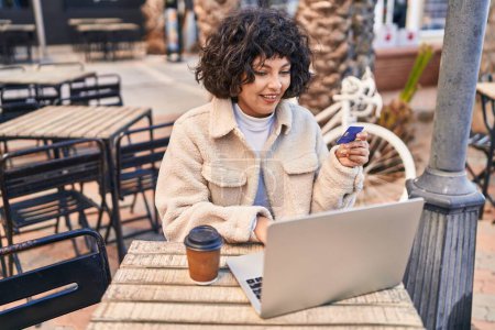 Foto de Joven mujer hispana hermosa usando portátil y tarjeta de crédito sentado en la mesa en la terraza de la cafetería - Imagen libre de derechos