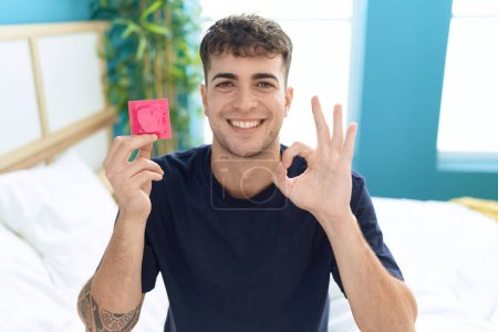 Téléchargez les photos : Jeune homme hispanique assis sur le lit tenant préservatif faisant signe ok avec les doigts, sourire geste amical excellent symbole - en image libre de droit