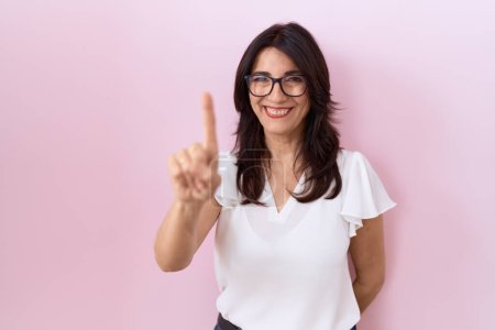 Foto de Mujer hispana de mediana edad con camiseta blanca casual y gafas que muestran y señalan con el dedo número uno mientras sonríe confiada y feliz. - Imagen libre de derechos
