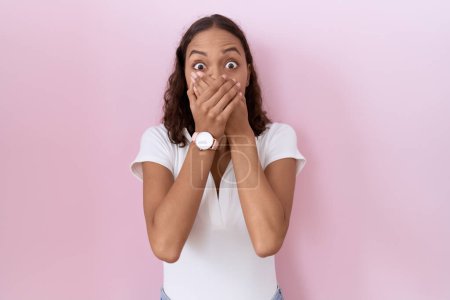 Foto de Mujer hispana joven que llevaba una camiseta blanca casual sorprendió cubriendo la boca con las manos por error. concepto secreto. - Imagen libre de derechos