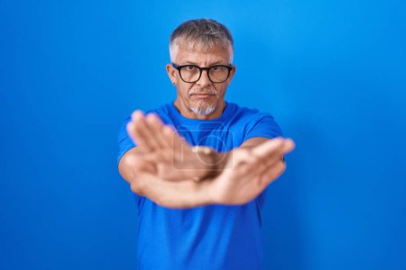 Foto de Hombre hispano con el pelo gris de pie sobre fondo azul expresión de rechazo cruzando brazos y palmas haciendo signo negativo, cara enojada - Imagen libre de derechos