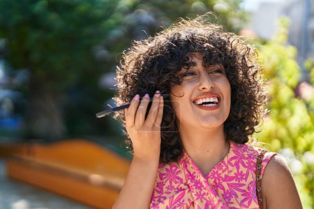 Téléchargez les photos : Jeune femme du Moyen-Orient souriant confiant écoute message audio par le smartphone au parc - en image libre de droit