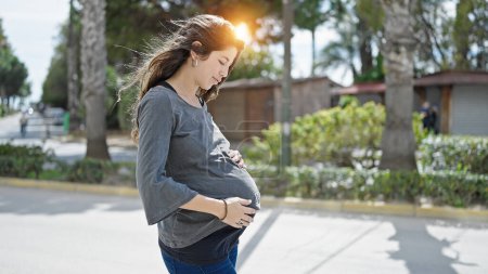 Foto de Mujer embarazada joven de pie con expresión seria tocando el vientre en el parque - Imagen libre de derechos