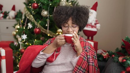 Téléchargez les photos : Jeune femme afro-américaine buvant du café assis sur le canapé près de l'arbre de Noël à la maison - en image libre de droit