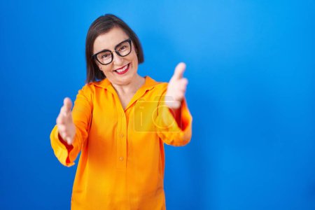 Téléchargez les photos : Femme hispanique du Moyen Âge portant des lunettes debout sur fond bleu regardant la caméra souriant à bras ouverts pour un câlin. expression joyeuse embrassant le bonheur. - en image libre de droit