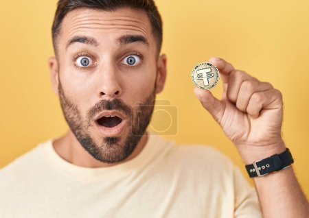 Foto de Hombre hispano guapo sosteniendo la moneda criptomoneda atado asustado y sorprendido con la boca abierta para sorpresa, cara de incredulidad - Imagen libre de derechos