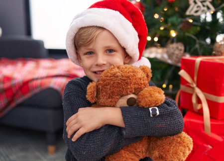Foto de Adorable niño abrazando oso de peluche de pie junto al árbol de Navidad en casa - Imagen libre de derechos