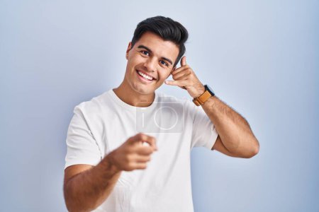 Foto de Hombre hispano de pie sobre fondo azul sonriendo haciendo hablar por teléfono y señalándote. Llámame.. - Imagen libre de derechos